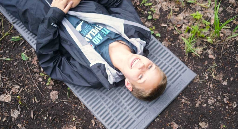 a person lays on a sleeping pad on the ground a smiles at the camera